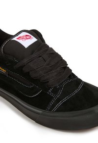 Sneaker in suede nera con linguetta in rete nera, lacci spessi neri, cuciture bianche, suola in gomma e un patch con logo rosso e bianco sulla linguetta.