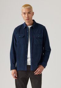Marinblaues Hemd mit Knopfleiste aus weichem Stoff, ausgestattet mit zwei Brusttaschen und einem klassischen Kragen. Über einem weißen Shirt getragen.