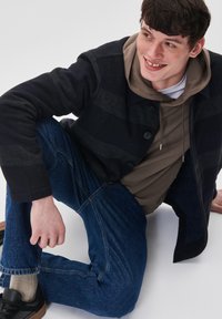 Chaqueta a rayas multicolores en azul marino y gris sobre una sudadera beige, combinada con jeans azules y zapatillas negras. Conjunto casual, materiales texturizados.