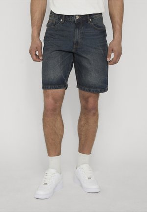 Homme portant un short en jean bleu foncé, des chaussettes blanches et des baskets blanches, debout devant un fond uni gris clair.