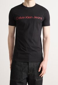 Černé bavlněné tričko s červeným textem "Calvin Klein Jeans" na hrudi. Klasický kulatý výstřih a přiléhavé krátké rukávy.
