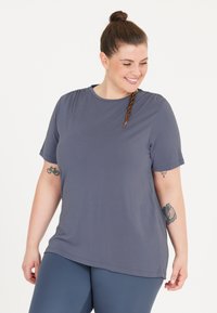 Grå kortærmet t-shirt med rund hals, glat tekstur og subtile samlingdetaljer ved skuldrene. Bæret med blå leggings.