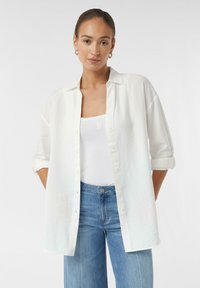 White oversized button-up shirt with rolled sleeves, worn over a white tank top, paired with light blue jeans. Smooth texture, casual design.
