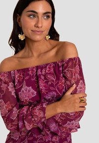 Schoudervrije paarse jurk met bloemmotieven, doorzichtige mouwen en rimpelranden. Model draagt gouden oorbellen en een gouden ring.