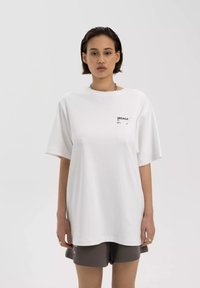 Witte oversized t-shirt met korte mouwen, voorzien van een zwart logo op de linkerborst. De stof lijkt zacht en soepel.