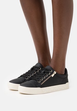 Sorte lave sneakers med hvide såler, snørebånd og sidelåse, båret på mørkhudede ben mod en ensfarvet hvid baggrund.