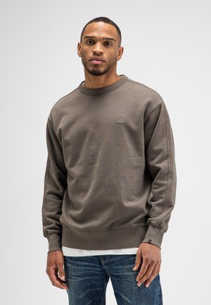 Man wearing a brown crewneck sweatshirt over a white shirt and blue jeans, standing against a plain light background.