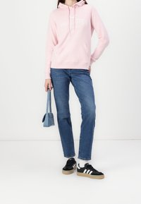 Osoba v růžovém mikině Hollister, modrých džínách a černých teniskách Adidas, držící malou modrou kabelku na bílém pozadí.