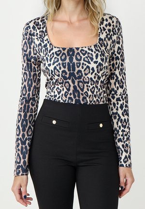 Vrouw draagt een top met lange mouwen in luipaardprint en een vierkante hals, ingestopt in een zwarte broek met hoge taille en gulp met knoopzakken.