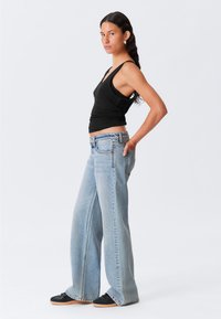 Svart ärmlös topp med en enskilt passform, matchad med ljusblå vida jeans och svarta avslappnade skor. Modellen står i profil.