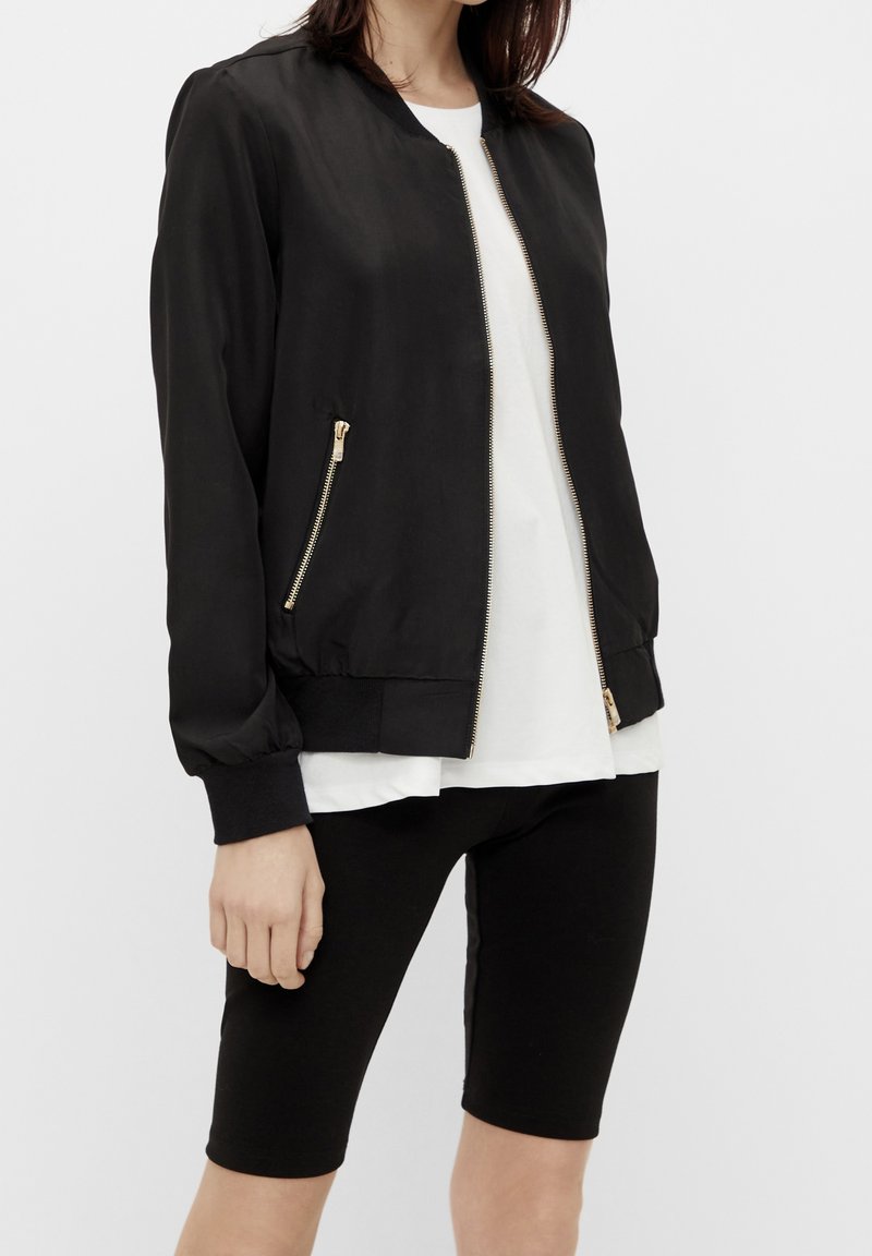 Mujer vestida con una chaqueta bomber negra con cremalleras doradas sobre una camisa blanca y pantalones cortos de bicicleta negros hasta la rodilla, de pie contra un fondo blanco.