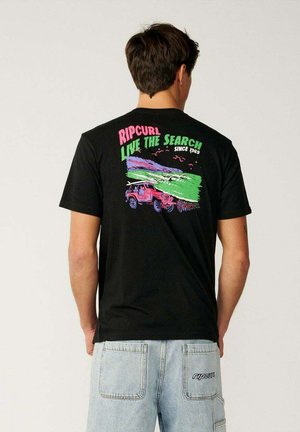 Homme portant un t-shirt noir avec un motif coloré Rip Curl "Live the Search" et un jean bleu clair, vu de dos.