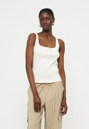 Kvinde iført hvid, ærmeløs ribstrikket top med bindedetalje foran og beige cargobukser, stående mod en ensfarvet lys baggrund.