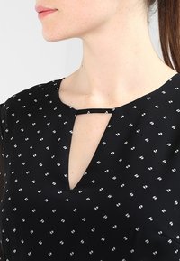 Blouse noire avec une petite ouverture en forme de clé au niveau du col, ornée d'un motif de petits pois blancs. Texture lisse, manches courtes.