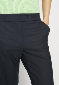 Gros plan d'une personne portant un pantalon noir habillé avec la main dans la poche et un haut en maille texturée vert clair rentré à l'intérieur.