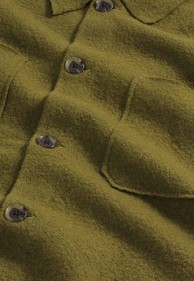 Camicia in lana verde oliva, testurizzata, con abbottonatura frontale, due tasche sul petto e grandi bottoni rotondi. Tessuto morbido con una superficie leggermente pelosa.