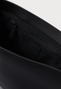 Juodos odos krepšio vidus su užtrauktuku uždaru skyriumi ir siūtu prekės ženklo ženklu viduje, priešais paprastą šviesų foną.