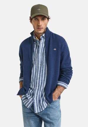 Dunkelblauer Reißverschluss-Pullover über einem hellblauen und weißen gestreiften Hemd, kombiniert mit hellblauen Jeans und einer olivgrünen Mütze.