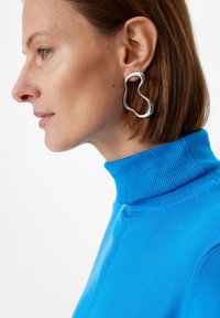 Srebrne abstrakcyjne kolczyki z falistym kształtem, uzupełnione niebieskim swetrem typu golf. Gładka, odblaskowa tekstura metalu.