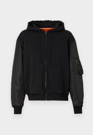 Μαύρο φούτερ με φερμουάρ, με λείο ύφασμα στο σώμα και μανίκια που μοιάζουν με σατέν. Διαθέτει εμπρόσθια τσέπη και επιπλέον τσέπη με φερμουάρ στο μανίκι.