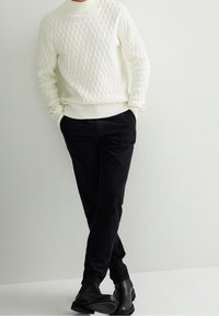 Osoba v bílém texturovaném svetru, černých manšestrových kalhotách a černých botách, stojící s rukama v kapsách na čistém bílém pozadí.