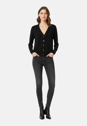 Donna con cardigan nero abbottonato, jeans skinny grigio scuro e stivaletti neri, in piedi con le mani nelle tasche su sfondo bianco.