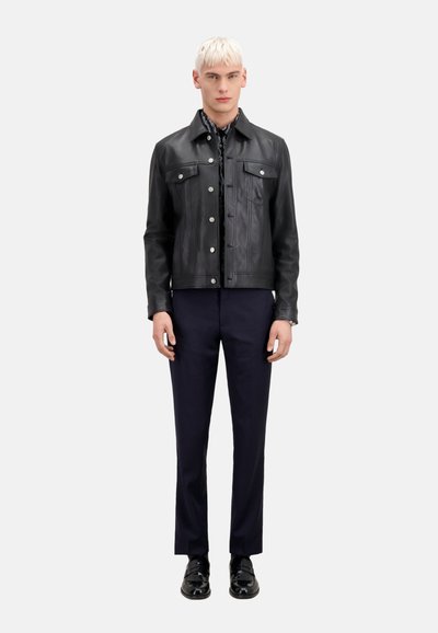 Giubbotto di pelle nera con chiusura a bottoni, due tasche al petto, colletto in stile denim, abbinato a pantaloni blu scuro e scarpe lucide.