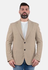 Blazer beige con texture, due tasche frontali, chiusura a un bottone e revers a scollatura profonda, indossato sopra una camicia bianca e jeans blu.