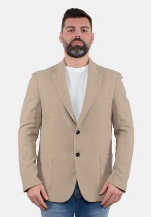 Blazer beige con texture, due tasche frontali, chiusura a un bottone e revers a scollatura profonda, indossato sopra una camicia bianca e jeans blu.