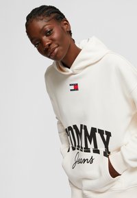 Felpa leggera beige realizzata in tessuto morbido, con un grande logo nero "TOMMY" e un'etichetta piccola rossa e blu sul petto, dotata di una tasca anteriore.