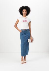 Hvit t-skjorte med kirsebærgrafikk, kombinert med et høyhåndtalt, denim maxi-skjørt. Modellen holder en liten, lys håndveske.