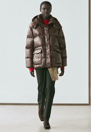 Bruine pufferjas met capuchon, voorzien van grote zakken en een glanzende textuur. Draagt over een rood shirt en donkere broek, aangevuld met bruine laarzen.