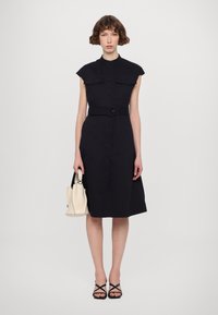 Crna košulja haljina s crnim remenom i kratkim rukavima, koja sadrži dva džepa na prsima i dugme za zatvaranje. Uparena s malom krem torbicom i crnim sandalama s trakicama.