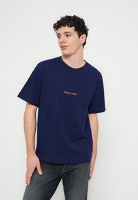 Nuori mies kiharatukkaisena, pukeutunut tummansiniseen T-paitaan, jossa keskellä rintaa oranssia tekstiä, ja tummanharmaisiin farkkuihin, seisoo valkoista taustaa vasten.