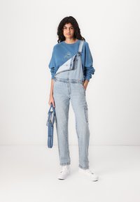 Pantalones de mezclilla en azul claro con una tirante bajado, combinados con un sudadera azul con un gráfico. Zapatillas blancas y una bolsa azul.