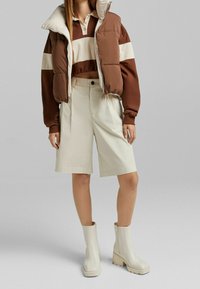 Gilet imbottito marrone sopra una maglietta a maniche lunghe a righe marrone e bianco, abbinato a pantaloni corti beige e stivali alla caviglia bianchi con tacco grosso.