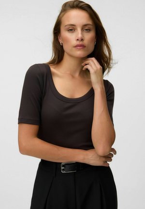 Jeune femme aux cheveux châtain clair portant un haut marron foncé à manches courtes et un pantalon noir avec ceinture, posant avec une main près du menton.