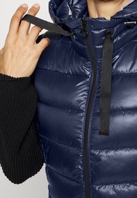 Personne ajustant un cordon noir sur une doudoune matelassée bleu marine portée sur un vêtement à manche en tricot noir.