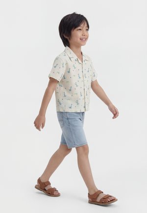 Ung gutt som går, iført en kortermet t-skjorte med dinosaurtrykk, lyse denimshorts og brune sandaler mot en ensfarget hvit bakgrunn.