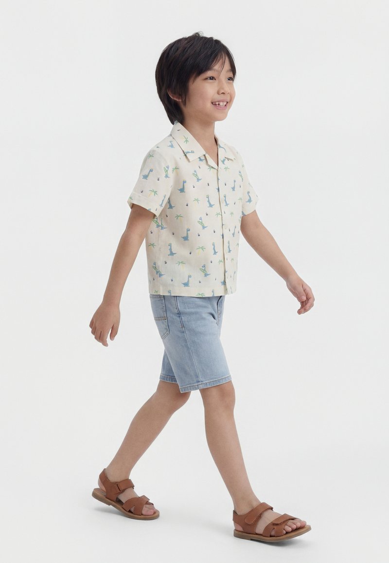 Mladi dječak hoda, nosi majicu s kratkim rukavima s dinosaurom, svijetle traper šortse i smeđe sandale na jednobojnoj bijeloj pozadini.