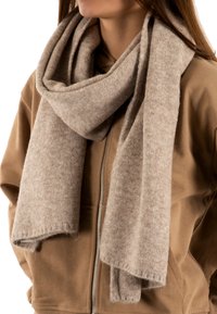 Sciarpa di lana beige con una texture morbida, appesa in modo lasco attorno al collo, caratterizzata da un design semplice e bordi sfrangiati, abbinata a una felpa marrone con zip.