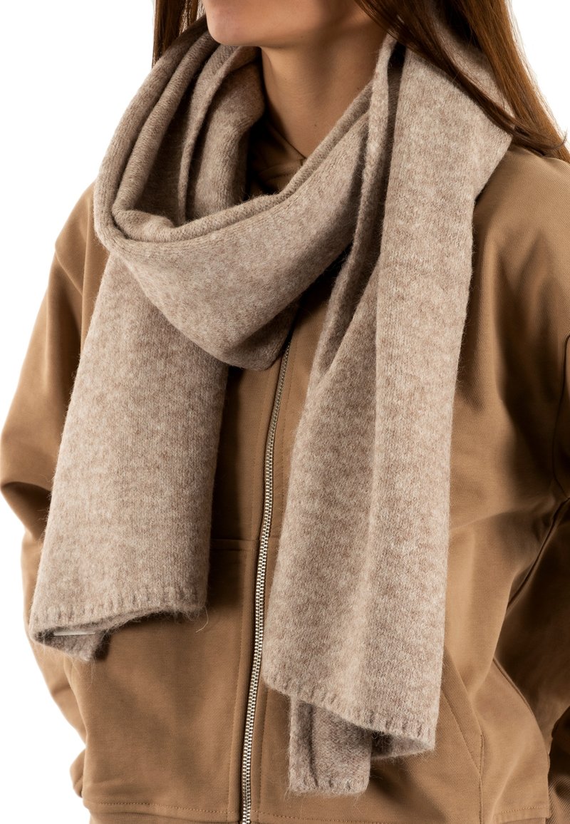 Sciarpa di lana beige con una texture morbida, appesa in modo lasco attorno al collo, caratterizzata da un design semplice e bordi sfrangiati, abbinata a una felpa marrone con zip.