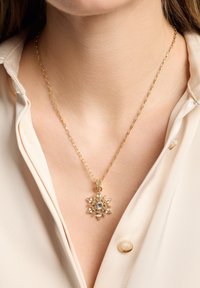 Gul kædehalssmykke med en snefnugpendant pyntet med klare rhinsten og en central ædelsten. Båret over en lysefarvet skjorte.