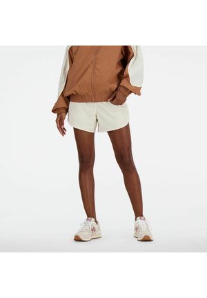 Person iført en brun og hvid jakke med lyse beige shorts og beige sneakers, stående på en ensfarvet hvid baggrund.