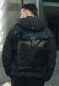 Black leather backpack with a roll-top design and silver hardware, worn over a black puffer jacket. Features a adjustable strap.