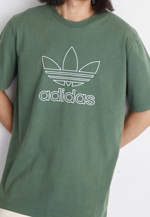 Persoon draagt een groen Adidas T-shirt met wit trefoil-logo en "adidas" tekst op de borst tegen een effen achtergrond.