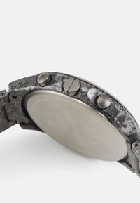 Montre analogique avec un bracelet texturé en noir et gris, dos en acier inoxydable et deux boutons poussoirs métalliques sur le côté.