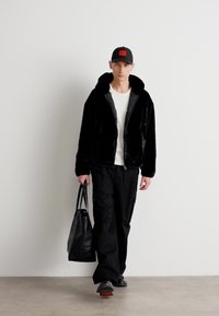 Black faux fur jacket with a hood, white t-shirt, black cargo pants, and black sneakers. Carries a black tote bag and wears a cap.