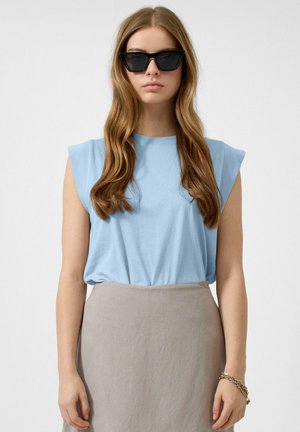 Junge Frau mit langen, welligen Haaren, die schwarze Cat-Eye-Sonnenbrillen, ein hellblaues ärmelloses Oberteil, einen beigen Rock und ein goldenes Armband trägt.