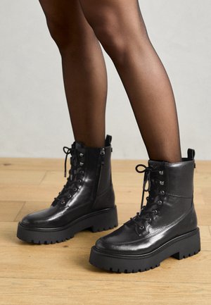 Crne kožnečizme do gležnja s debelim gumenim potplatom, vezanjem sprijeda i bočnim patentnim zatvaračem. Imaju teksturiranu površinu i izražen uzorak potplata.